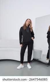 Bright studio interior features a young blonde woman posing confidently in a black hoodie and pants. She wears white sneakers, showcasing a contemporary fashion style suitable for casual outings.