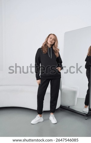 Bright studio interior features a young blonde woman posing confidently in a black hoodie and pants. She wears white sneakers, showcasing a contemporary fashion style suitable for casual outings.