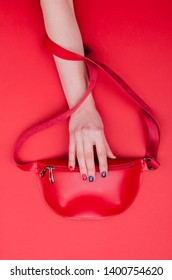 Bright Summer Fashion Ladies Accessories. Stylish Red Leather Handbag Or Fanny Pack On A Red Background. Women Hand With Bright Manicure Hold Bag From Top. Top View. Flat Lay. Copy Space. Mock Up