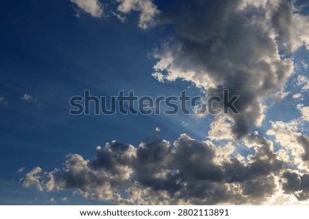 Bright sun rays break through fluffy clouds against a rich blue sky backdrop. The picturesque skyscape showcases a beautiful play of light and shadow during a sunny day.