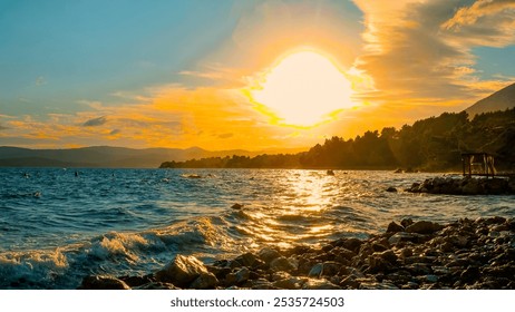 Bright sun setting over the ocean, casting golden light across the waves and rocky shore. Scenic coastal landscape with silhouetted trees and distant mountains under a glowing sky. - Powered by Shutterstock