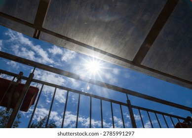 A bright sun shining through a translucent roof structure, resembling a terrace or balcony with a metal railing.