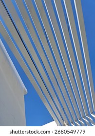 The bright sunlight casts sharp shadows beneath each slat, highlighting the depth and three-dimensionality of the structure. The stark white of the beams against the deep blue creates a powerful visua