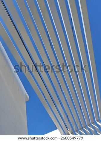 The bright sunlight casts sharp shadows beneath each slat, highlighting the depth and three-dimensionality of the structure. The stark white of the beams against the deep blue creates a powerful visua