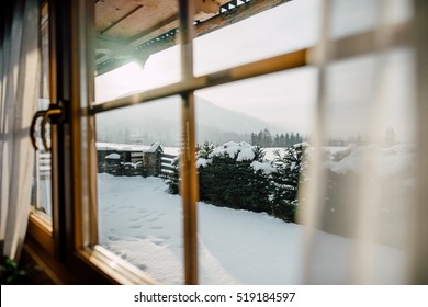 Bright Sunlight Shines Through The Wooden Cottage House Window. Beautiful Snowy Winter Morning Behind The Chalet Window. Sunshine And Sparkling Snow On Fir Trees In The Cabin Backyard.