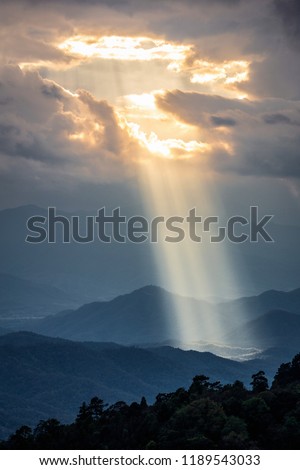 Bright sunlight shining through hole of clouds to dark scene of mountain range before sunset in Thailand rainforest area.