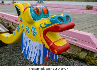 A bright and vibrant painted dragon's head from a dragon boat competition or race. The asian and chinese traditional vessel is near a barge on a sandy shore. The dragon has yellow and blue on its head - Shutterstock ID 1161458977