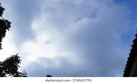 Bright white clouds float in a clear blue midday sky, with the shadow of tree branches framing the view of a house roof. This serene scene combines natural elements and architectural details, perfect for design projects, backgrounds, or visual storytelling