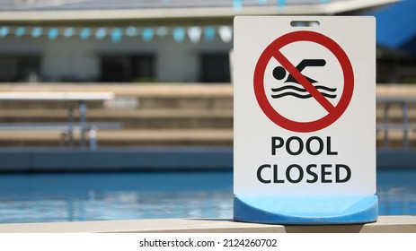 A bright white sign at a local outdoor pool facility saying pool is closed. No entry to water. Clear clean blue water at public baths and no swimming symbol.  - Shutterstock ID 2124260702