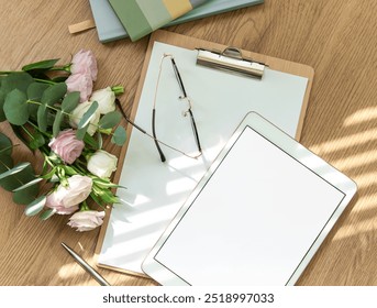 A bright workspace features a blank tablet, an open notebook, stationery tools, and fresh flowers arranged on a wooden table, inviting creativity and planning. - Powered by Shutterstock