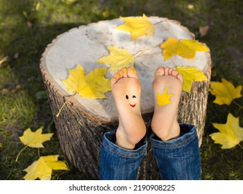 Bright Yellow Autumn Leaves On A Stump. Painted Smile On Bare Feet Toddler Child. Joy, Cheerful, Positive Thinking, Happy Childhood. Hello, Autumn. Seasonal Fun Creative Ideas