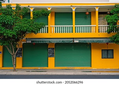 Bright yellow building facade with green roller shutters, front view.  Green trees on the sides. Traditional Asian architecture, colonial style.  Vietnam, Nha Trang: 2020-10-23
