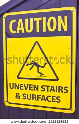 Bright yellow caution sign with a triangular symbol, warning passersby of uneven stairs and surfaces. Bold black text and a silhouette figure in a falling motion emphasize the warning to ensure safety