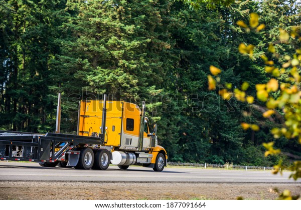 Bright yellow long hauler big rig red industrial\
semi truck transporting empty step down semi trailer running on the\
straight highway road with green trees on the side to warehouse for\
next load