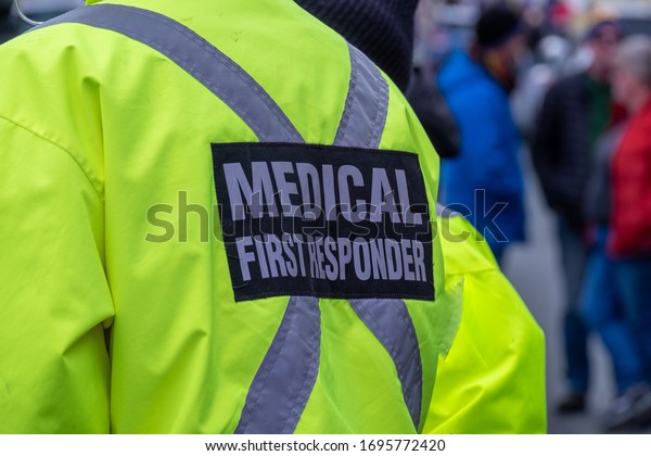 A bright yellow medical first responder uniform\
being worn by a large male in a crowded street.The coat has a grey\
reflective cross across the back of the emt or ent. There are\
people in the distance.