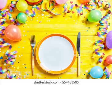 Bright Yellow Place Setting For The Carnival Season With Coiled Streamers, Party Balloons And Confetti Around A Clean Empty Plate With Knife And Fork Viewed From Above