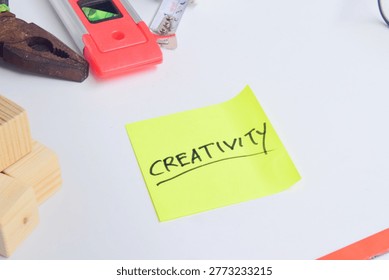 A bright yellow sticky note with 'CREATIVITY' handwritten in black ink, placed on a white surface among various tools and building blocks. Evokes ideas of inspiration, planning, and problem-solving.