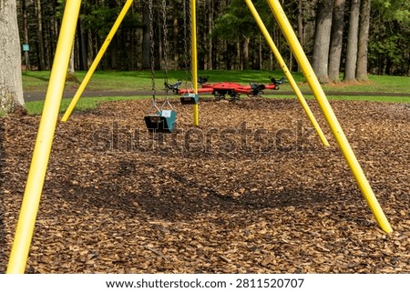 A bright yellow swing set with two empty swings and a red spinner is set up on a surface of wood chips in a park with a forest background