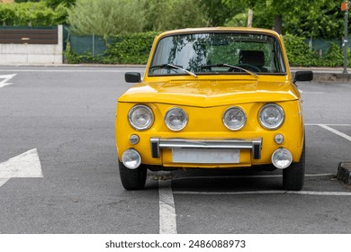 A bright yellow vintage car with classic round headlights is parked slightly crooked in an empty parking space on a quiet street surrounded by greenery and trees. - Powered by Shutterstock