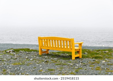 A bright yellow wooden bench with armrest on a rocky beach overlooking a foggy view of the ocean. There's grass scattered and growing under the seat. The cloudy sky and shore are grey and misty. - Powered by Shutterstock