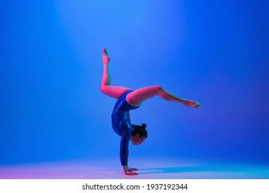 Bright. Young flexible girl isolated on blue studio background in neon light. Young female model practicing artistic gymnastics. Exercises for flexibility, balance. Grace in motion, sport, action.