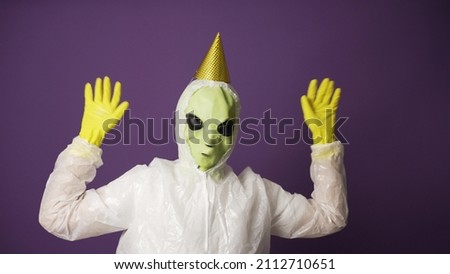 Bright-green alien with big black eye sockets wearing protection costume and gloves with birthday cap dances against violet wall closeup