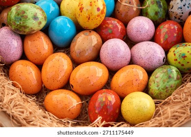 Brightly colored ceramic Easter eggs with speckled patterns lie together in a straw-filled basket, creating a cheerful spring and holiday decoration.