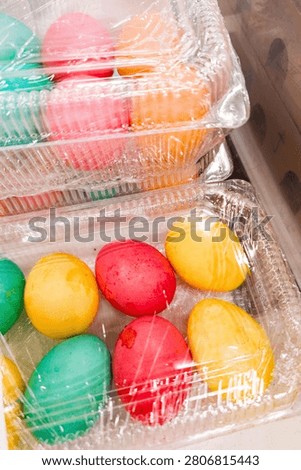 Brightly colored Easter eggs in shades of red, yellow, green, and orange are arranged inside clear plastic containers covered with plastic wrap. The vibrant hues create a festive and cheerful appearan