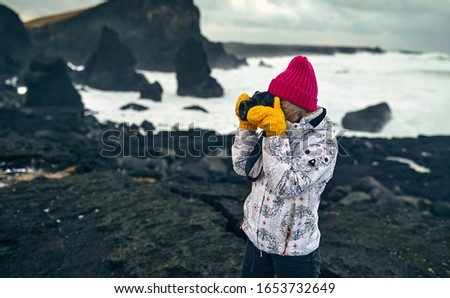 Brightly dressed young girl tourist makes a photo of a beautiful seascape, standing on the shore with black volcanic sand in cloudy weather. Travel, leisure, tourism.