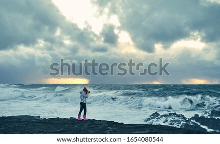 Brightly dressed young girl tourist makes a photo of a beautiful seascape, standing on the shore with black volcanic sand in cloudy weather. Travel, leisure, tourism.