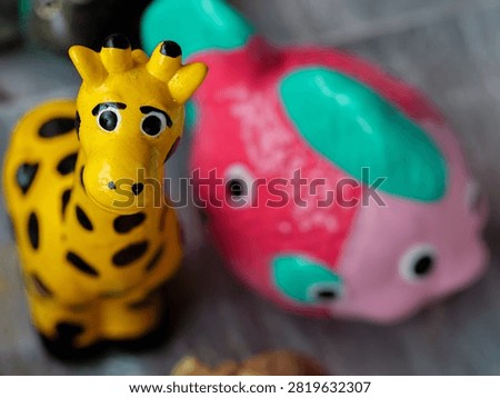A brightly lit close-up captures two whimsical ceramic animal figurines: a yellow giraffe with black spots looking towards the viewer, and a pink and teal fish slightly out of focus behind it. Their playful design evokes a sense of childhood joy and imagination