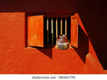 A brightly painted red wall and window of a village home in state of Maharashtra, India with a 'kalashi' or 'ghagar' which is a copper vessel used to store potable water.