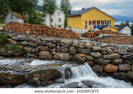 A brightly painted yellow house, a large stack of chopped logs and a wall made of stone to keep the rapids of a flowing river on path make for a beautiful Norwegian landscape during a cloudy day. 
