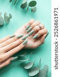 Brightly polished long green nails are elegantly arranged on a mint backdrop, accentuated by scattered eucalyptus leaves for a fresh, natural look.