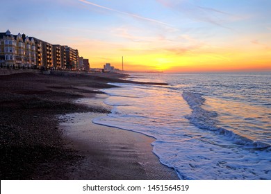 Brighton beach and seafront at sunset, the sun is is orange and low in the sky setting just behind Brighton pier casting an orange glow in the sky, the waves are breaking in the foreground, Brighton