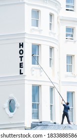 Brighton, Sussex / UK - 9 June 2019: A Window Cleaner Uses A Very Long Brush - Hose Combination To Clean Glass On Upper Floors. The Man Stands On A Lead Roof Of A White Building Pointing To 'HOTEL'.