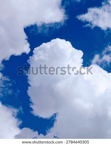 A brilliant blue sky forms the background for a massive, brilliant white cumulus cloud formation. The central cloud boasts a remarkably dense, fluffy texture with soft, rounded edges that stand out sharply against the deep blue atmosphere. Its towering shape creates a striking sense of vertical depth, catching the sunlight perfectly to emphasize its bright white surfaces and subtle grayish shadows. Whispy fragments of smaller clouds frame the main structure, adding movement to the scene. Captured from a low angle looking straight up, the composition evokes a peaceful, awe-inspiring feeling of looking into the vastness of nature.