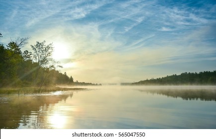 Brilliant and bright mid-summer sunrise on a lake.   Warm water and cooler air at daybreak creates misty fog patches.  Still water along a calm, quiet Ontario lakeside.