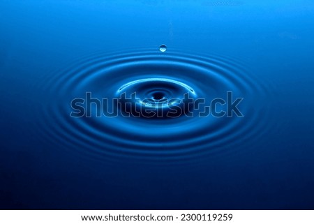 A brilliant rich blue pool of water gently rippled with concentric circles with a single drop of floating spherical water above.