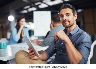 Bringing digital ideas to the meeting. Portrait of a man sitting at a table in an office using a digital tablet with colleagues working in the background. - Shutterstock ID 2129942822