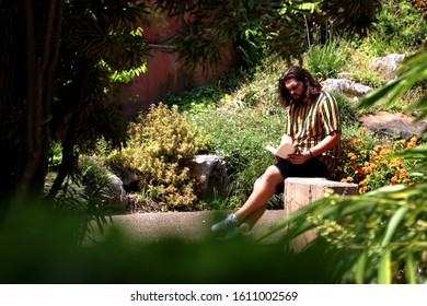 Brisbane QLD Australia January 09/2020 - An Adult Male In His Early 20's Sits Reading A Book In A Park Among The Trees, Bushes And Flowers On A Warm Summer Day.