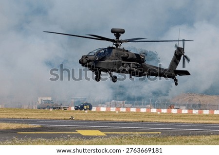 A British Army Apache attack helicopter hovers low over an airfield, generating a large cloud of dust and exhaust during a maneuver.