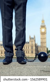 British Politician With Ball And Chain Shackled To His Ankle Stands In Front Of The Houses Of Parliament At Westminster, London, UK