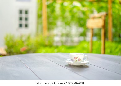 British Tea Cup Style With Drawing Pink Flower Pattern On It, On The Wood Dark Brown Table In The Morning With Blur Garden And Window.