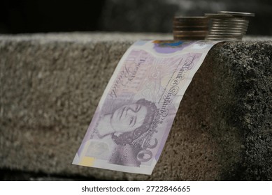 A British twenty-pound note drapes elegantly over a textured concrete edge, accompanied by coin stacks, against a dark, undefined background, suggesting financial flow.