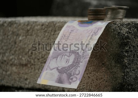 A British twenty-pound note drapes elegantly over a textured concrete edge, accompanied by coin stacks, against a dark, undefined background, suggesting financial flow.