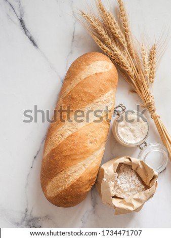 British White Bloomer or European sourdough Baton loaf bread on white marble background. Fresh loaf bread and glass jar with sourdough starter, flour in paper bag, ears. Top view. Copy space. Vertical