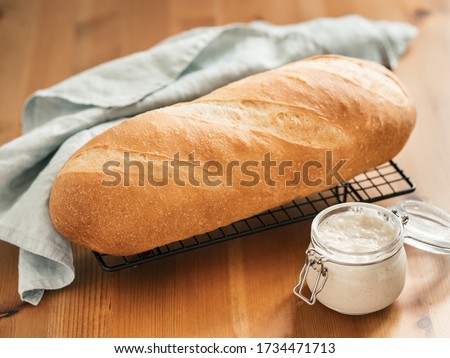 British White Bloomer or European sourdough Baton loaf bread on wooden background. Fresh loaf bread and glass jar with sourdough starter. Copy space