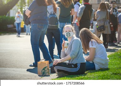 Brno, Czech Republic - April 23 2020: A Girl Who Drinks Starbucks Coffee Under A Cloth Face Mask With The Starbucks Logo, Sits On The Ground Next To A Queue Of People For A Corona Virus Test.