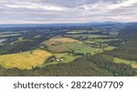Broad aerial panorama of patchwork fields and meadows across rolling hills, with forest patches, winding roads, scattered villages, power lines, orchards, windmills, and cloudy sky